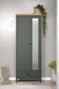 A tall, dark green wardrobe with a mirrored door, a wooden top, and a drawer at the bottom. It is placed against a light gray wall, with a woven basket and a rug in front of it. A plant in a basket is on top of the wardrobe, and another plant is in a pot t Podgląd zdjęcia produktu