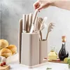 A hand is placing a knife into a kitchen utensil holder filled with knives and other kitchen tools. In the foreground, there are rolls of bread, cherry tomatoes, a cutting board, and bottles of oil and vinegar. Produktbild-Vorschau