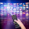 A hand holding a remote control in front of a wall of screens displaying various content. Podgląd zdjęcia produktu