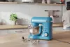 A blue stand mixer is on a wooden countertop in a kitchen, with various attachments laid out in front of it. A person is visible in the background. Produktbild-Vorschau