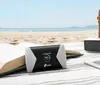 A mobile hotspot sits on a blanket on a beach, next to an open book and a speaker, with the ocean in the background. Produktbild-Vorschau