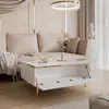 A modern living room scene featuring a white high-gloss coffee table with gold legs and open storage compartments, placed in front of a beige sofa. Produktbild-Vorschau