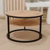 A round coffee table with a wood-look top and shelf, supported by a black metal frame, is shown in a room with light wood flooring and a slatted wooden partition in the background. Voir les images du produit