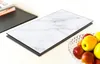 A glass cutting board with a marble pattern is placed on a white surface, next to a bowl of apples and a stack of books. Produktbild-Vorschau
