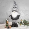 A decorative Christmas gnome figure is displayed on a white fluffy surface, with a glass vase containing decorative elements and a pine branch with ornaments in the background. Náhled obrázku produktu