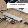 A silver USB hub with various ports and accessories connected to a laptop on a wooden desk. Náhled obrázku produktu
