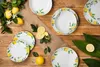 A collection of white porcelain plates with a lemon and leaf pattern, arranged on a wooden surface with lemons and green leaves. Anteprima dell’immagine prodotto