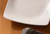A close-up shot of a white ceramic plate with a dark gray rock in the foreground, set against a brown background. Náhled obrázku produktu