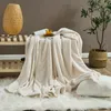 A cozy living room scene with a cream-colored blanket draped over a table, a decorative lamp, a plant in a basket, and a fluffy rug. Produktbild-Vorschau