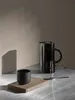 A black kettle and a black cup are placed on a marble surface, with a shadow cast on a tan wall. Produktbild-Vorschau