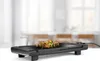 A black electric grill with food on it, placed on a white surface in front of a blurred background of a dining area. Anteprima dell’immagine prodotto