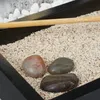 A close-up view of a miniature Zen garden with sand, stones, and a wooden rake. Náhled obrázku produktu