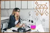 A woman is working at a desk with a computer and a camera. There are decorative diamond shapes overlaid on the image. Produktbild-Vorschau