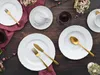 A table setting with white porcelain plates, cups, and gold cutlery, decorated with flowers. Produktbild-Vorschau