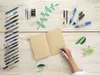 A hand is writing in an open notebook surrounded by various pens, pencils, and decorative leaves. Produktbild-Vorschau