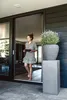 A woman is walking out of a doorway holding a tray, with a large potted plant to her right. Produktbild-Vorschau