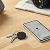 A Chipolo One Black Smart Key Locator is shown on a wooden surface next to keys and a smartphone displaying a map. Produktbild-Vorschau