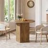 A round dining table with a fluted pedestal base is set with a vase of flowers, a glass of orange juice, and two apples. Two chairs are placed around the table, and a large clock hangs on the wall. Produktbild-Vorschau