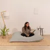 A woman is reading a book while sitting on a large, light gray beanbag chair in a minimalist room. Voir les images du produit