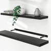 Two black floating shelves are shown mounted on a white wall. The top shelf has books, a white vase with a plant, and a grey vase. The bottom shelf is shown with its mounting brackets. Produktbild-Vorschau