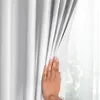 A hand is pulling back a white curtain to reveal a window with greenery outside. Produktbild-Vorschau