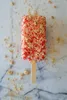 A single popsicle covered in red and tan crumbs on a marble surface. Produktbild-Vorschau