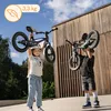 Two children are holding balance bikes above their heads in an outdoor setting with a wooden building in the background. Produktbild-Vorschau