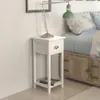 A white nightstand with a drawer and a shelf is placed next to a bed. Podgląd zdjęcia produktu