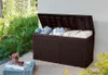 An open brown storage box filled with cushions and blankets, placed on a patio next to lush green plants. Podgląd zdjęcia produktu