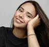 A young woman with freckles rests her head on her hand, showcasing a gold charm bracelet with a heart-shaped pendant on her wrist. Voir les images du produit