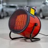 A red electric heater with a glowing heating element sits on a light-colored floor, with blurred cars in the background. Náhled obrázku produktu