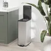 A stainless steel trash can with a foot pedal and open lid is shown next to a green cabinet and a large potted plant. Podgląd zdjęcia produktu
