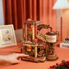 A 3D wooden puzzle of a gift factory is displayed on an orange surface, with a hand interacting with it. A framed picture and Christmas decorations are also visible. Podgląd zdjęcia produktu