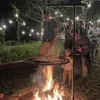 People gathered around a campfire at night, with string lights in the background. A grill is suspended over the fire. Podgląd zdjęcia produktu