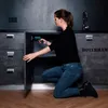 A woman is kneeling and using a cordless drill to assemble a cabinet. Produktbild-Vorschau