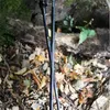 Two trekking poles are shown in a natural outdoor setting, resting against a fallen log with leaves and dirt on the ground. Podgląd zdjęcia produktu