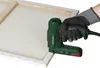 A hand wearing a glove is holding a green electric tacker and using it on the edge of a canvas. Produktbild-Vorschau
