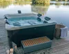 A hot tub is situated on a wooden deck next to a body of water, with a basket and blanket nearby. Náhľad obrázku produktu