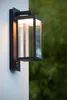 A modern black outdoor wall light with a glass enclosure is mounted on a textured white wall, with blurred green foliage in the background. Podgląd zdjęcia produktu