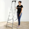 A woman stands next to a silver aluminum stepladder with 6 steps, on a wooden floor with a white wall in the background. Podgląd zdjęcia produktu