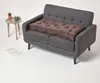 A brown tufted cushion is placed on a dark gray sofa. A small round side table with a cactus and a mug sits to the left of the sofa, and an open book lies on the floor. Náhled obrázku produktu