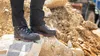 A person wearing black work pants and black work boots with pink accents stands on large rocks, with a pile of dirt and construction equipment in the background. Náhľad obrázku produktu