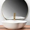 A white oval vessel sink with a gold faucet is shown in front of a round mirror. Produktbild-Vorschau