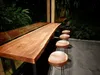 A long, live-edge wooden table with four bar stools in front of it, set against a backdrop of lush greenery. Produktbild-Vorschau