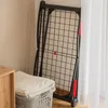 A black folding drying rack is leaning against a wall next to a window with white curtains. A laundry basket with a folded white towel is in the foreground. Produktbild-Vorschau
