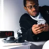 A young man in a bathrobe is lying in bed, looking at his phone. On the nightstand next to him are a digital alarm clock, a pair of headphones, and a book. Produktbild-Vorschau