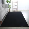 A black rug is laid out on a wooden floor in a living room setting, next to a sofa and a shelf with plants and books. Produktbild-Vorschau