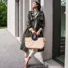 A woman stands next to a building, holding a shopper bag. Produktbild-Vorschau