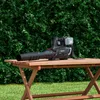 A cordless leaf blower rests on a wooden table in a garden setting, with a lush green hedge in the background. Produktbild-Vorschau