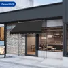 A black awning is extended over the entrance of a cafe or shop, with a brick wall on the left and a window showing the interior on the right. Náhľad obrázku produktu